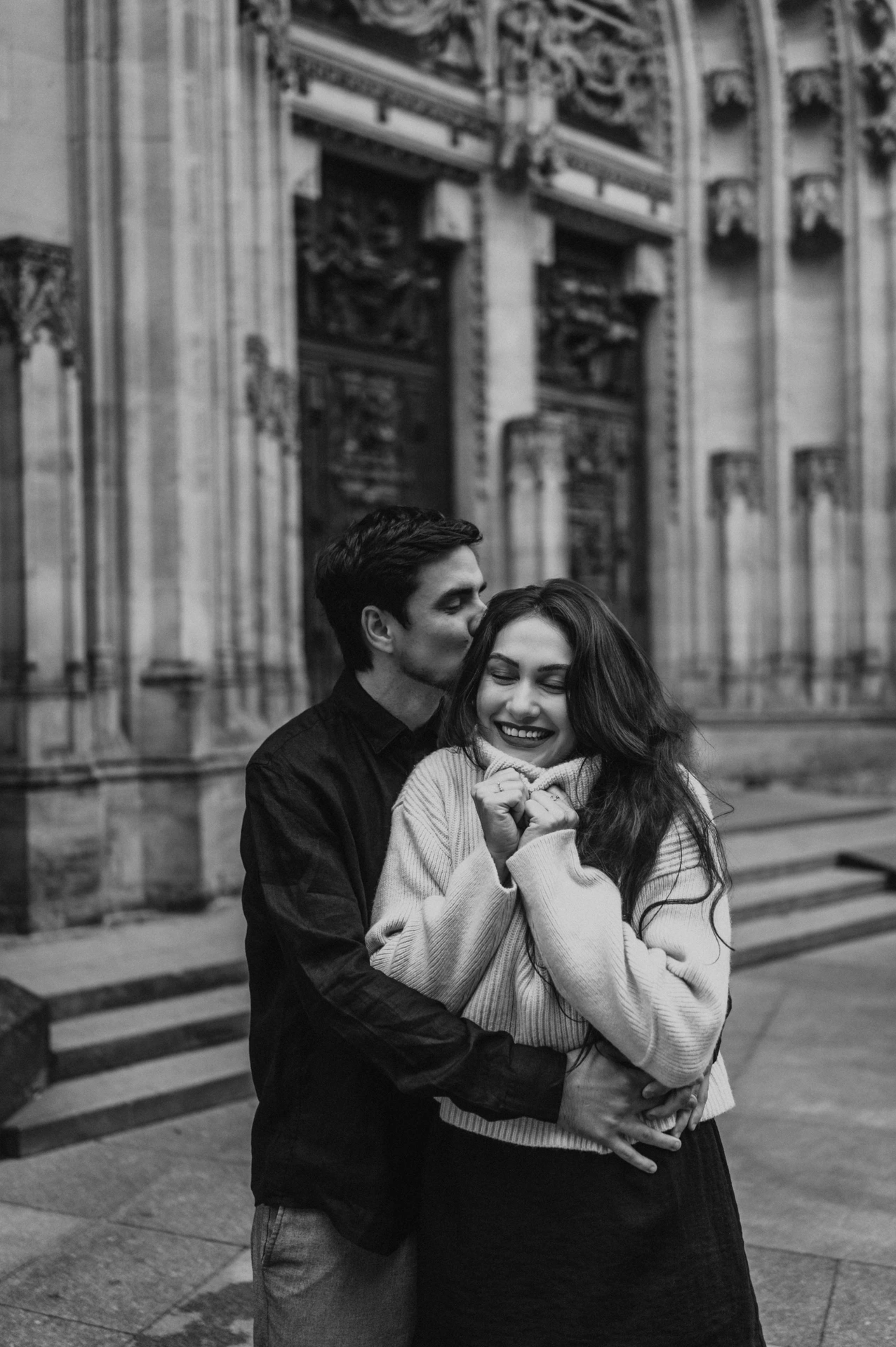 Black-and-white portrait of the woman smiling as her partner kisses her temple during their engagement photoshoot at Prague Castle.