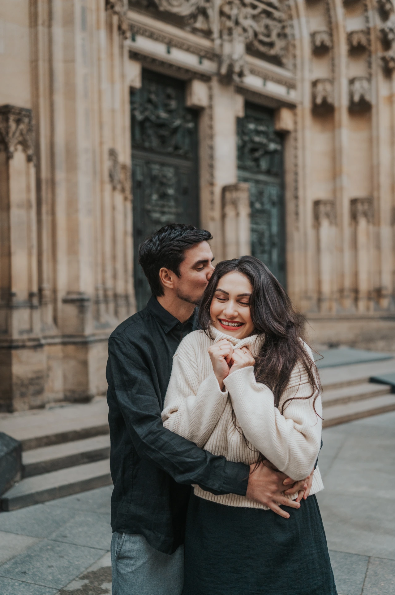 Prague engagement photoshoot with a temple kiss by St. Vitus Cathedral Woman smiling with her eyes closed as her partner kisses her temple beside St. Vitus Cathedral during their Prague engagement photoshoot.