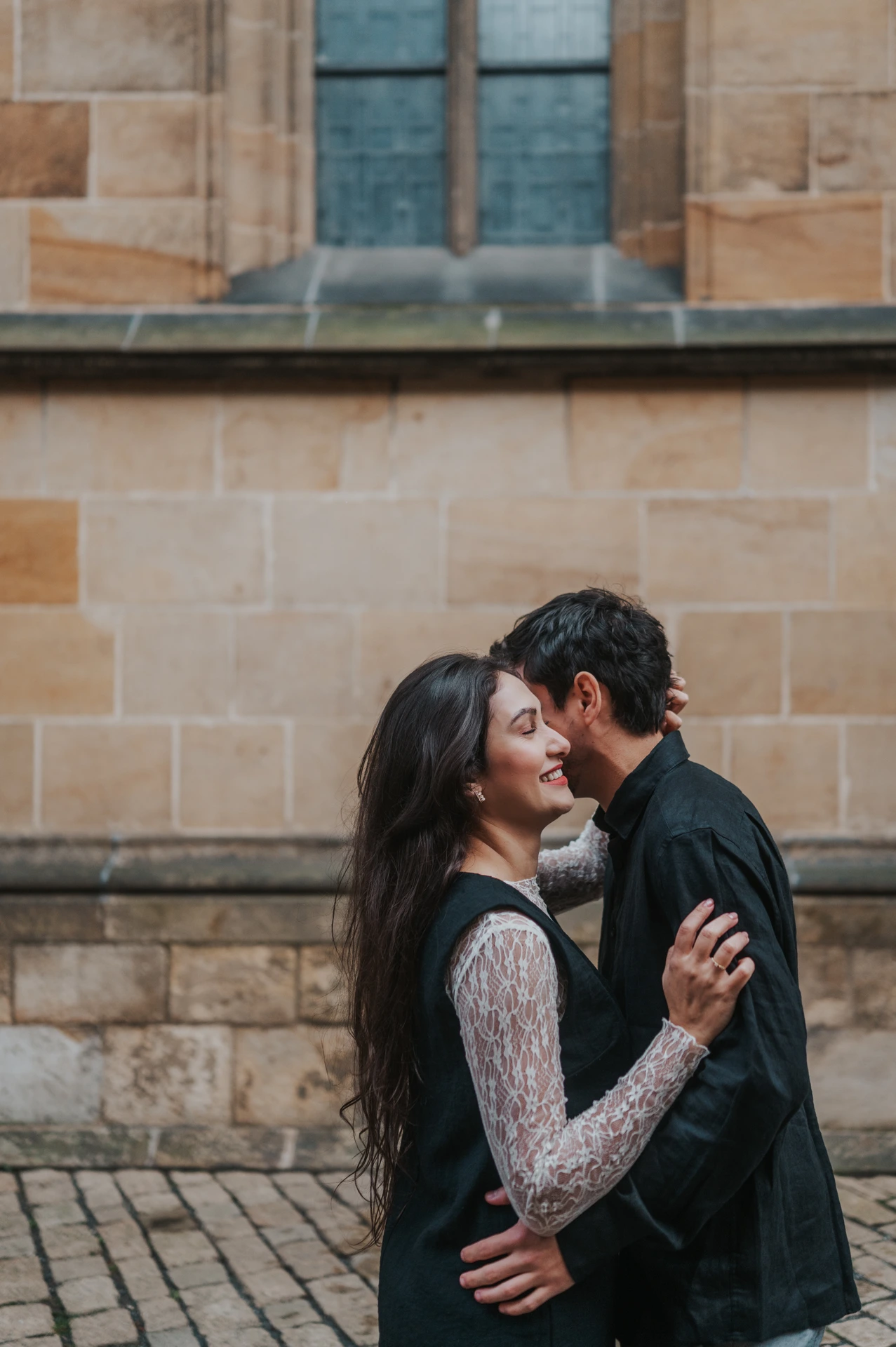 Couple holding each other close beside the stone cathedral wall during their engagement photoshoot in Prague.