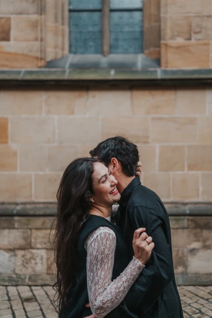 Prague engagement photoshoot with a kiss and a smile by the cathedral Woman smiling as her partner kisses the side of her head during their engagement photoshoot beside the cathedral wall in Prague.