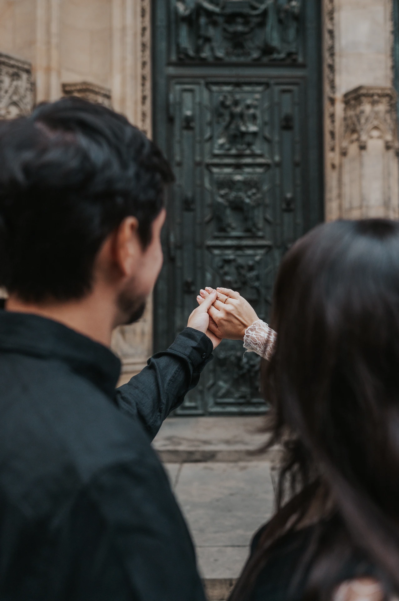 View over the couple's shoulders as they hold up their hands and engagement ring in front of the cathedral during their Prague photoshoot.
