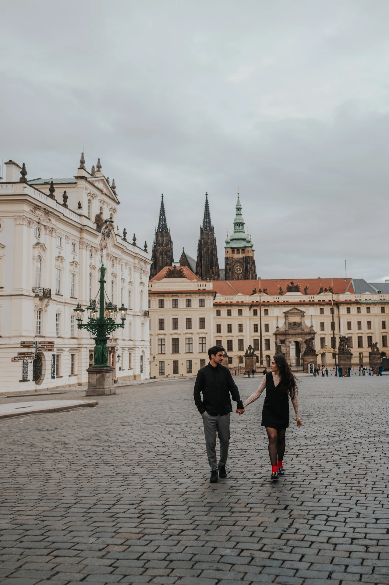 Couple walking together across the cobblestones of Hradcany Square with Prague Castle and cathedral spires behind them during their engagement photoshoot.