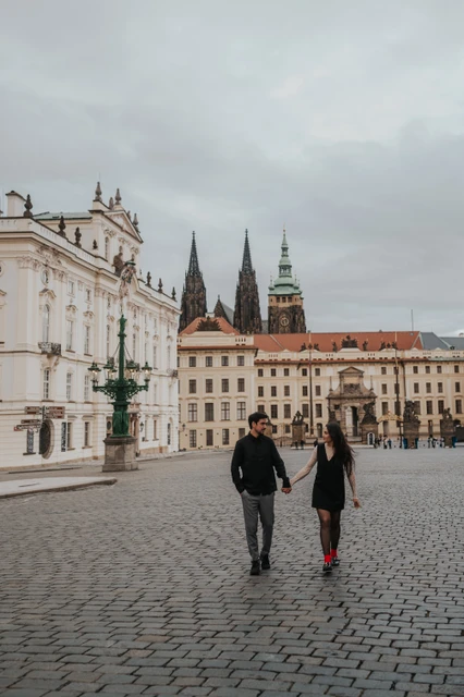 Prague engagement photoshoot walking across Hradcany Square Couple walking together across the cobblestones of Hradcany Square with Prague Castle and cathedral spires behind them during their engagement photoshoot.