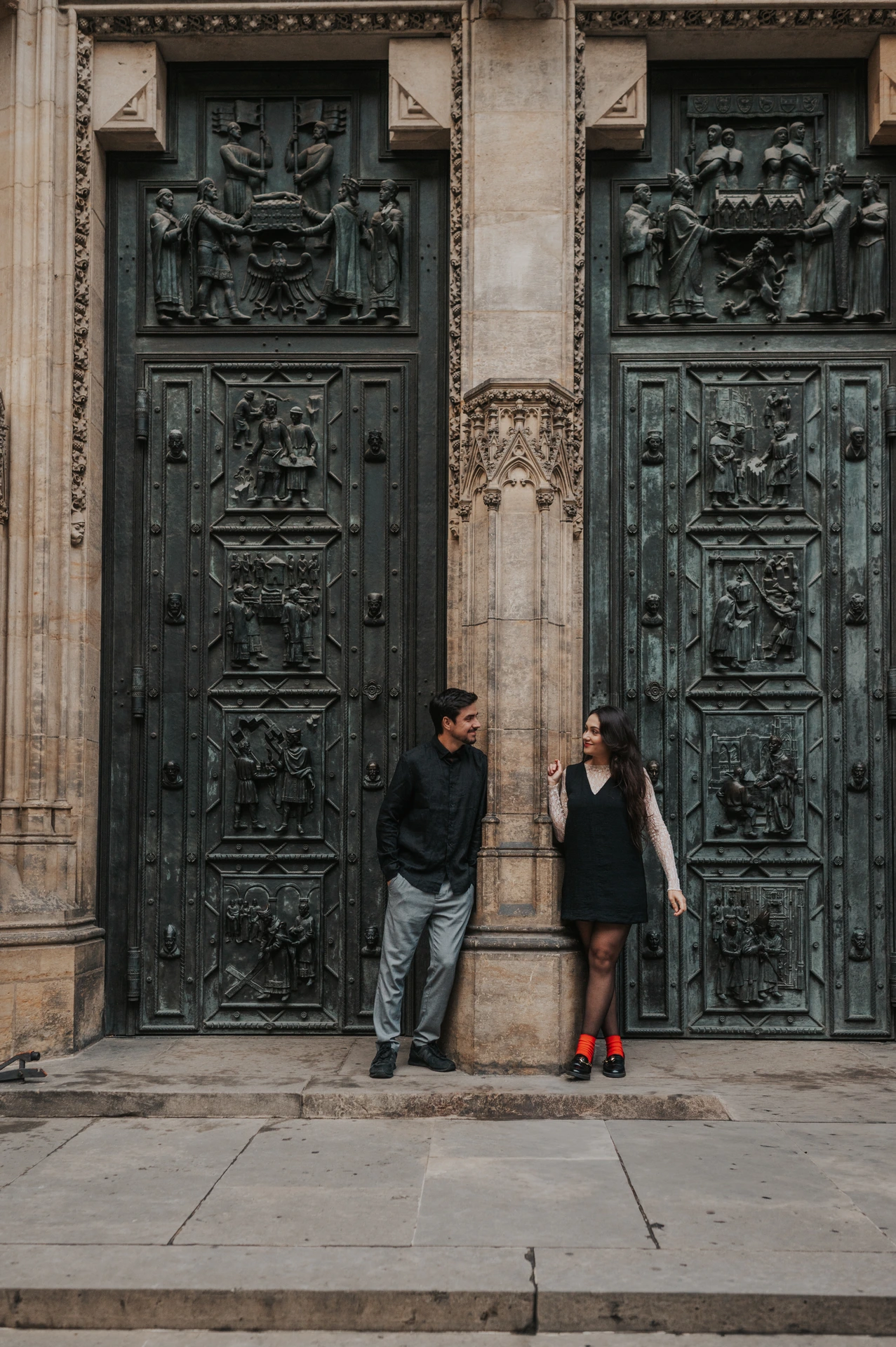 Couple standing either side of a stone column between the bronze St. Vitus Cathedral doors during their Prague engagement photoshoot.