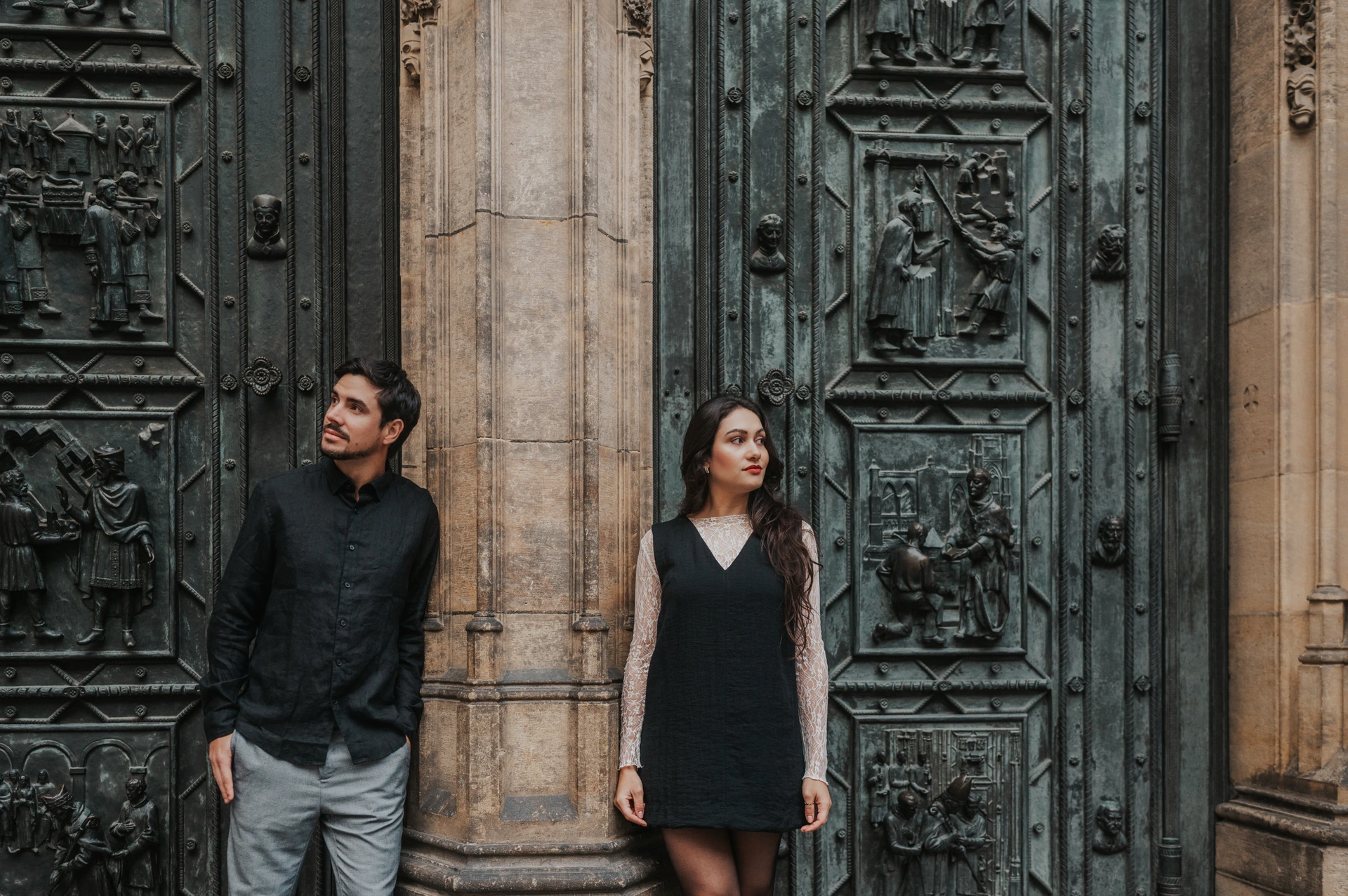 Landscape portrait of the couple looking in different directions beside the carved bronze cathedral doors during their engagement photoshoot in Prague.