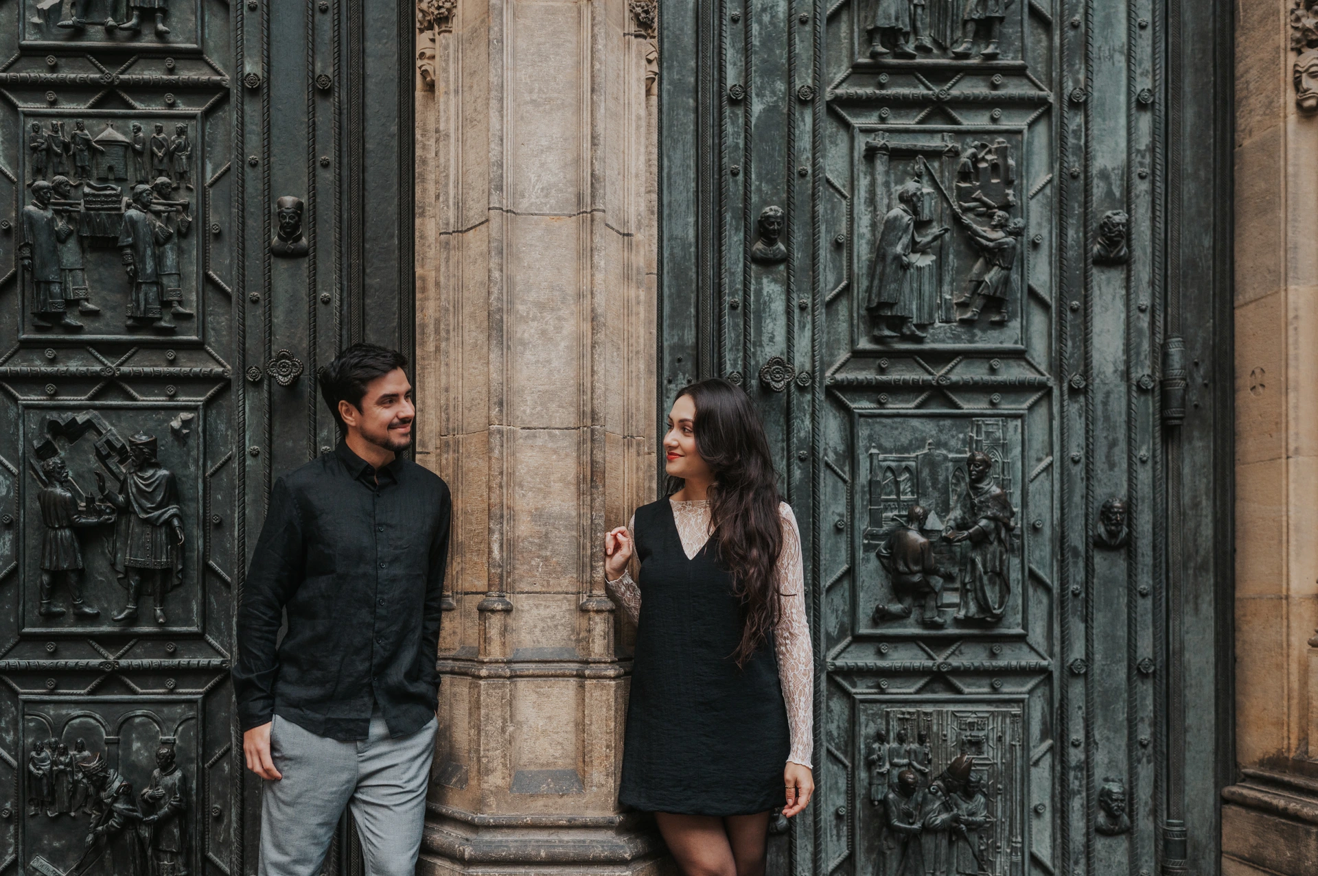 Close landscape portrait of the couple looking at each other between the bronze cathedral doors during their Prague engagement photoshoot.