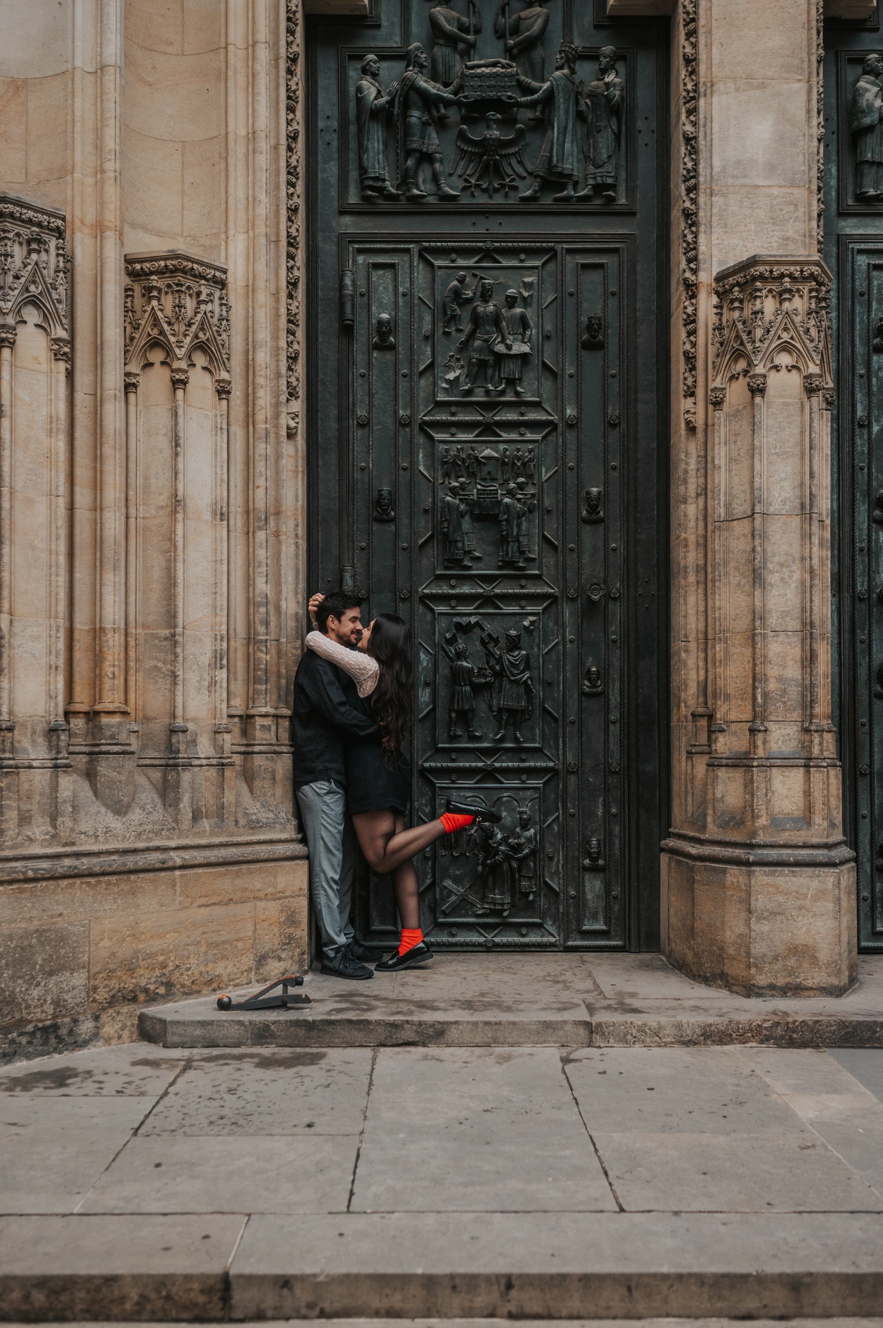 Couple kissing by the St. Vitus Cathedral doorway, with her red boot lifted behind her, during their engagement photoshoot in Prague.