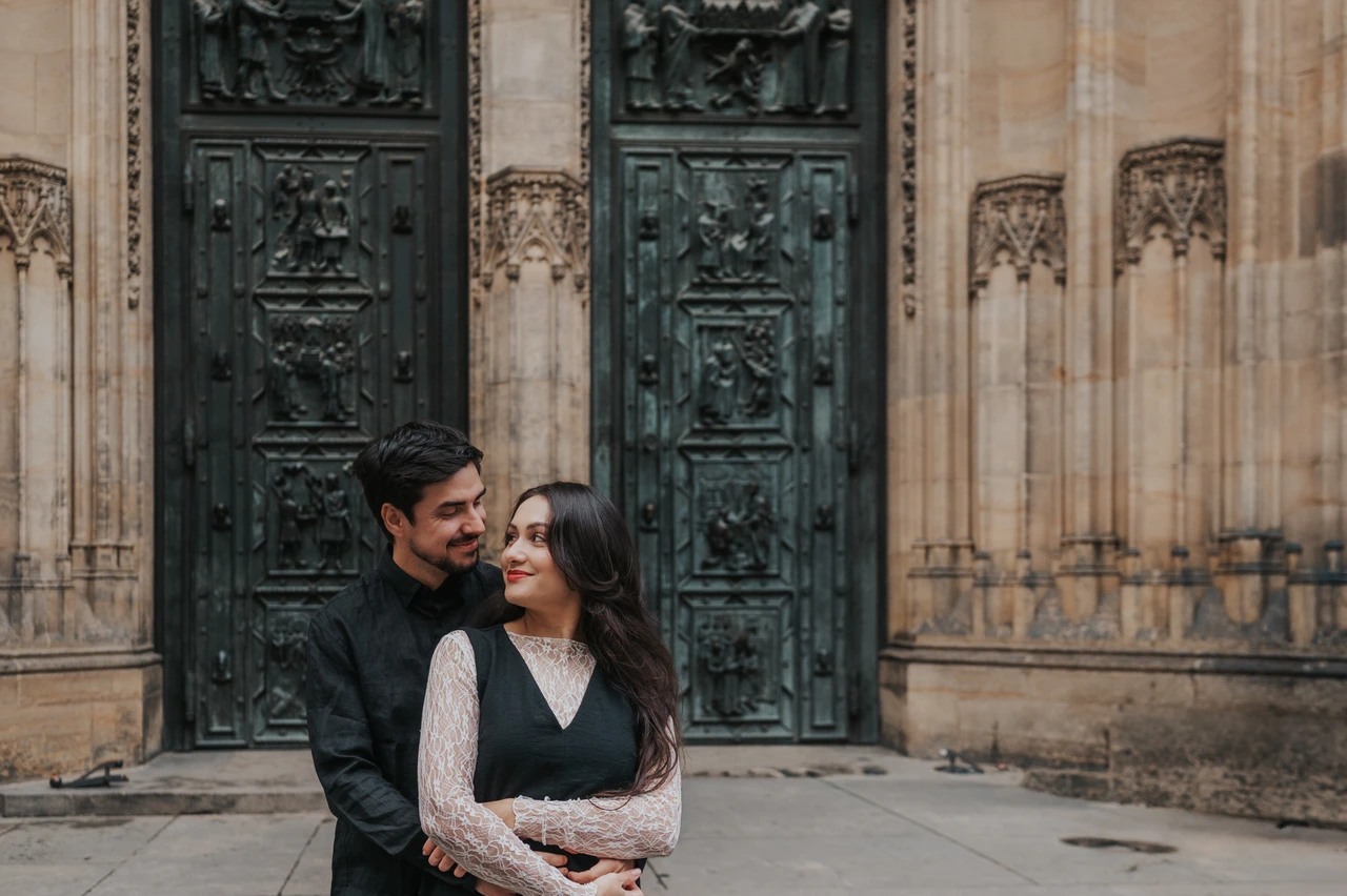 Prague engagement photoshoot with an embrace by the cathedral doors Landscape portrait of the couple smiling in an embrace with the cathedral doors behind them during their engagement photoshoot in Prague.