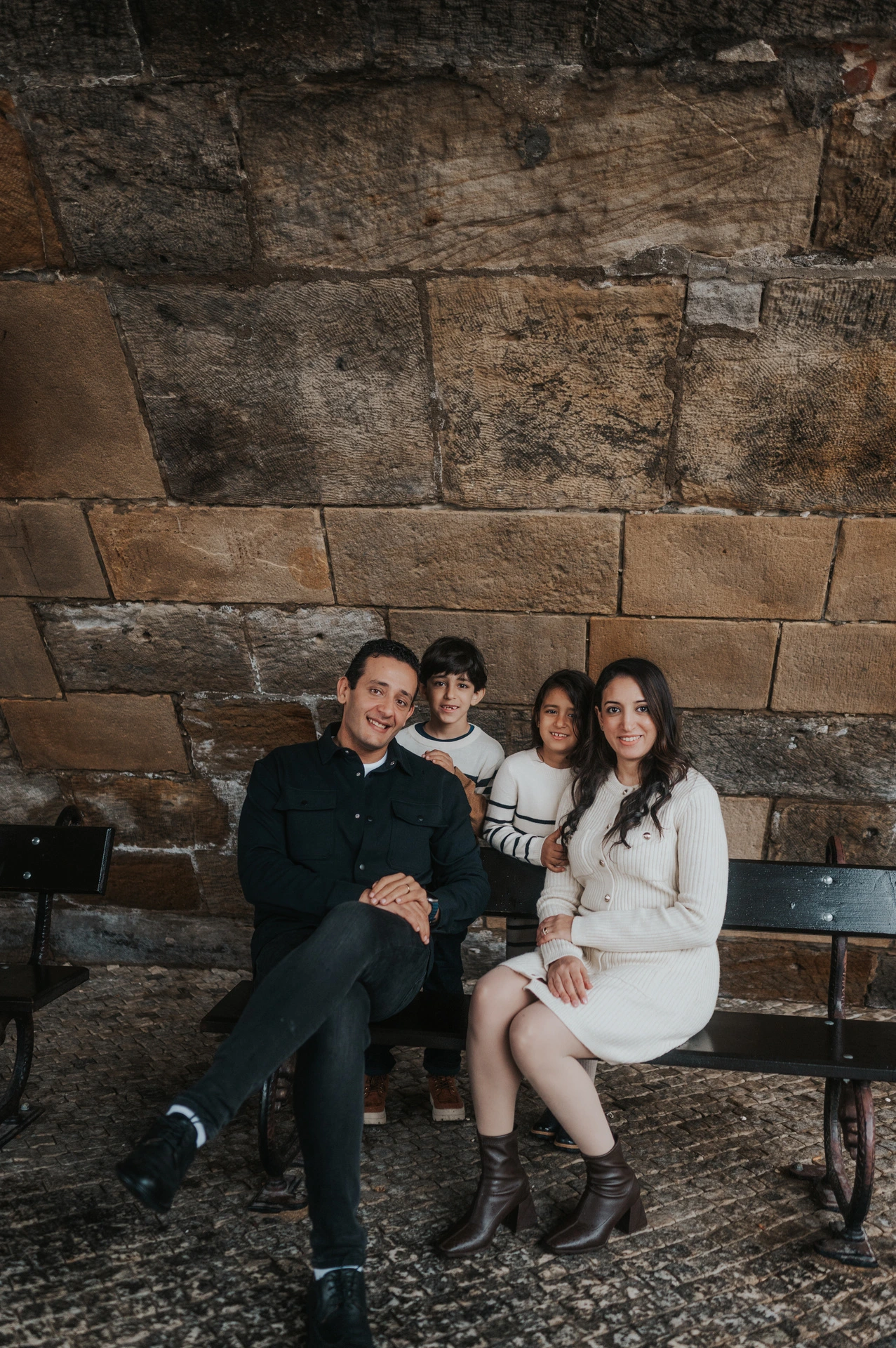 Parents and children smiling and leaning together on the bench below Charles Bridge during their Prague family photoshoot.