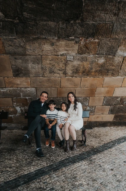 Prague family photoshoot: together on a bench beneath Charles Bridge Parents and their twins seated on a bench against the stonework beneath Charles Bridge during their Prague family photoshoot.