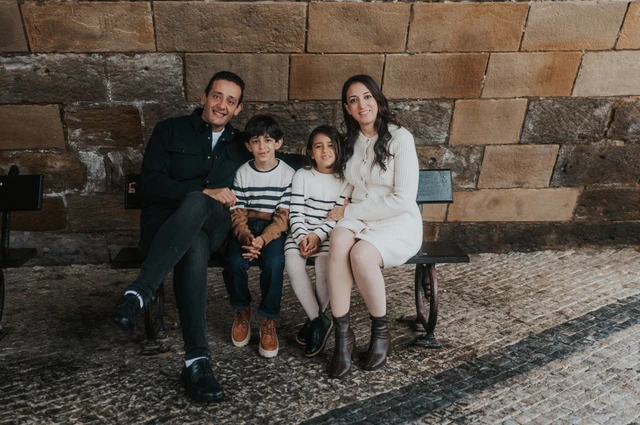 Wide portrait of the parents and their twins sitting closely together on a sheltered bench beneath Charles Bridge during their Prague family photoshoot.