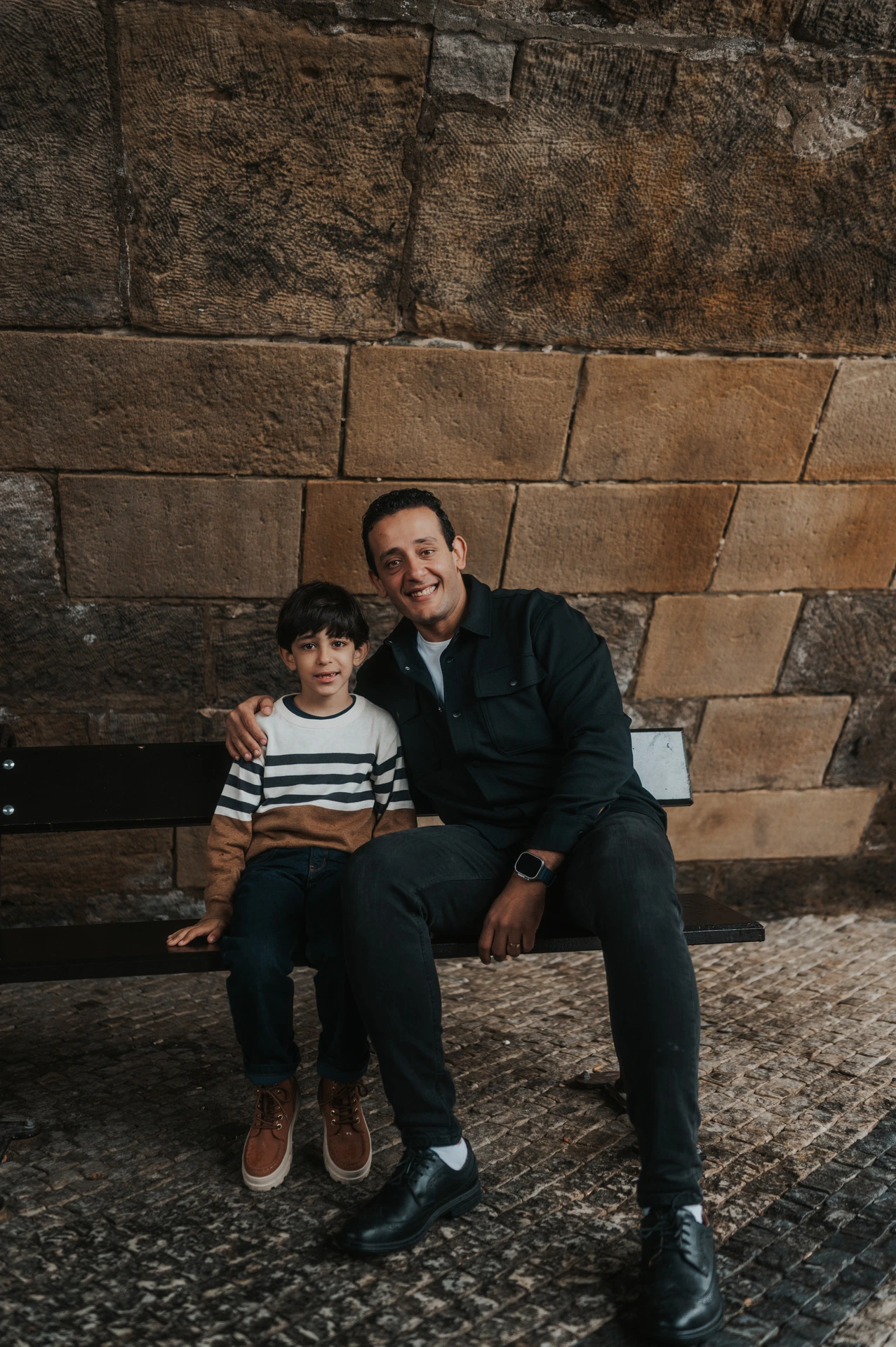 Father sitting beside his son with an arm around him on the sheltered bridge bench during their Prague family photoshoot.