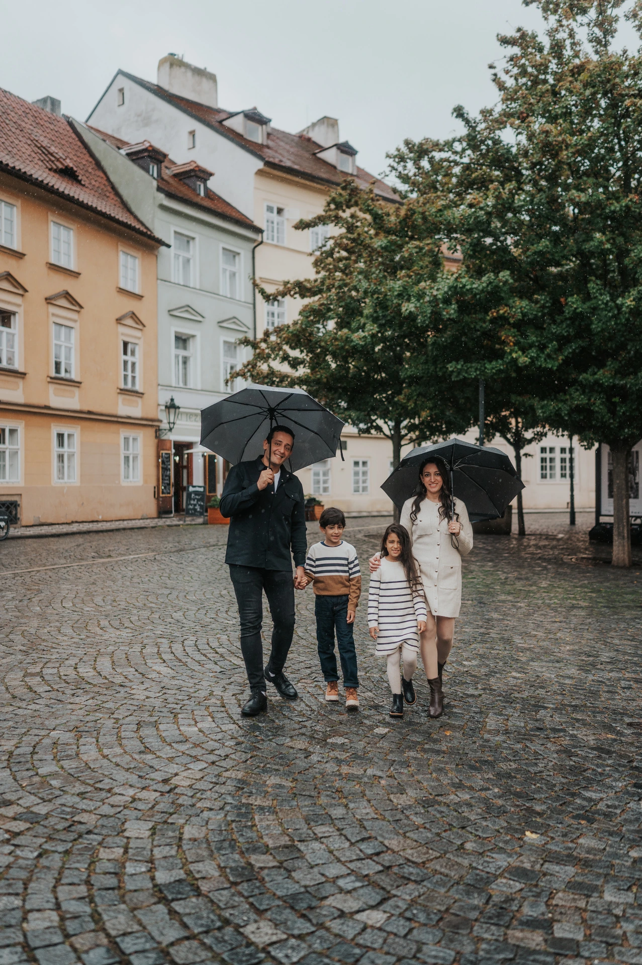 Family walking together with umbrellas across the wet cobblestones of Kampa during their Prague family photoshoot.