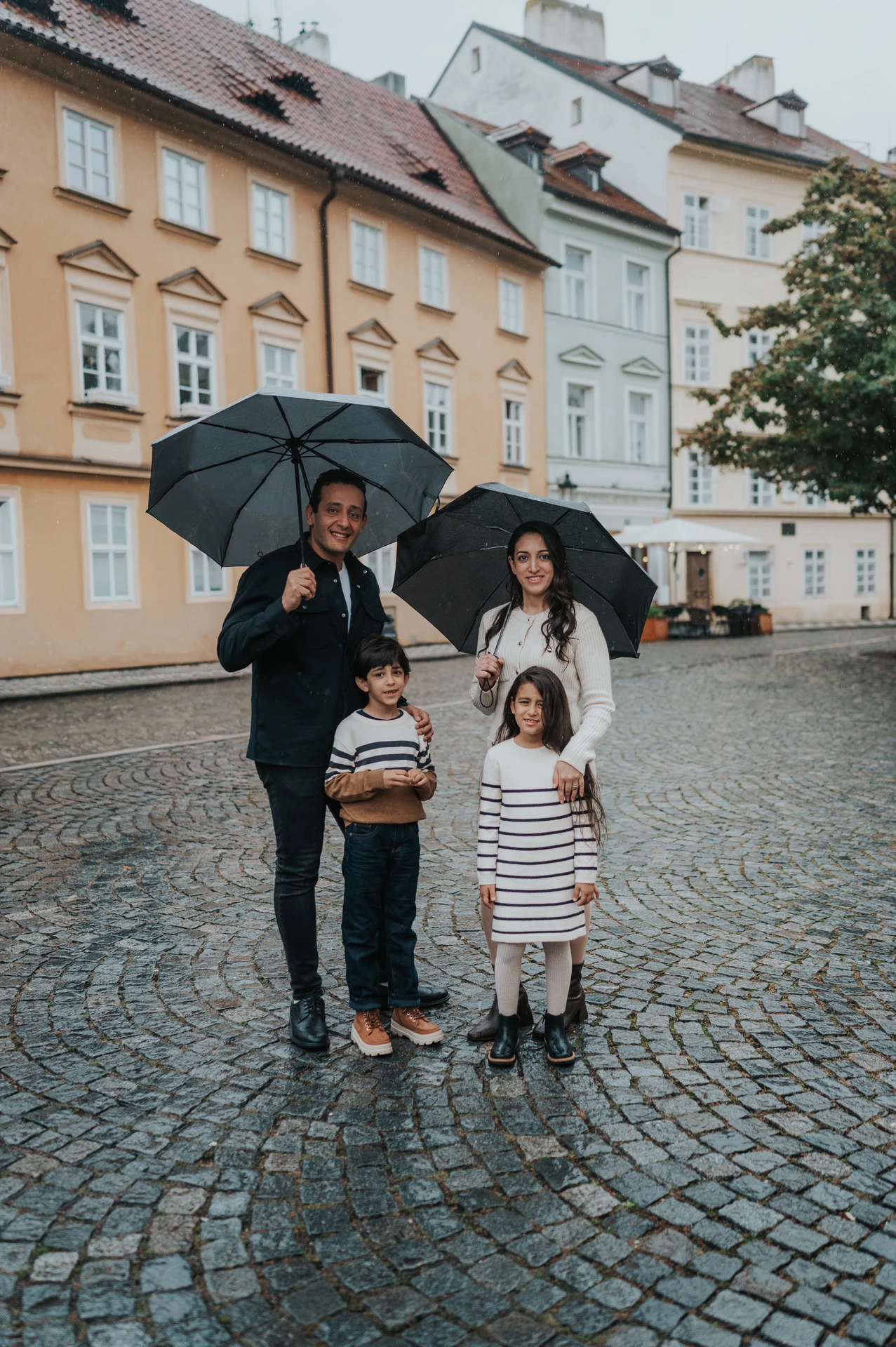 Parents and their twins standing together beneath umbrellas on wet cobblestones in Kampa during their Prague family photoshoot.