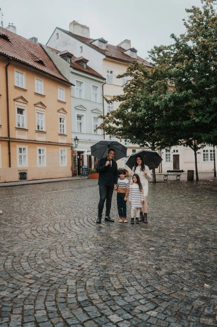 Prague family photoshoot: a rainy family portrait in Kampa Distant portrait of the family holding umbrellas in a Kampa square with pastel houses behind them during their Prague family photoshoot.