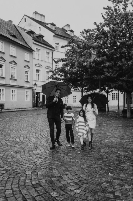 Prague family photoshoot: a black-and-white family walk in Kampa Black-and-white photograph of the family walking with umbrellas across a cobbled Kampa square during their Prague family photoshoot.