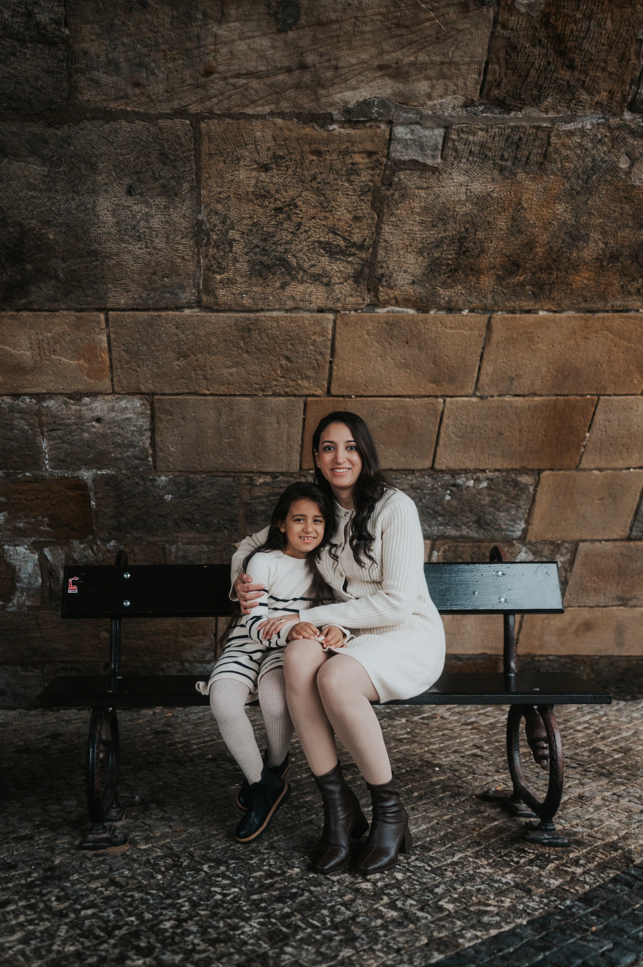 Mother and daughter sitting close together on a bench beneath Charles Bridge during their Prague family photoshoot.