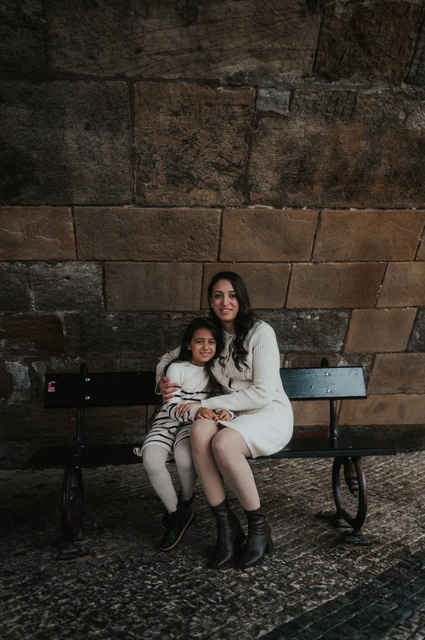 Prague family photoshoot: a mother-and-daughter cuddle under the bridge Mother embracing her daughter as they sit together on the sheltered bench during their Prague family photoshoot.