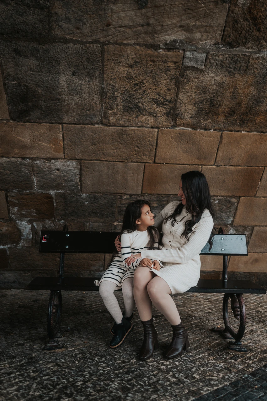 Prague family photoshoot: a shared look between mother and daughter Mother and daughter looking at each other while seated on the bench below Charles Bridge during their Prague family photoshoot.