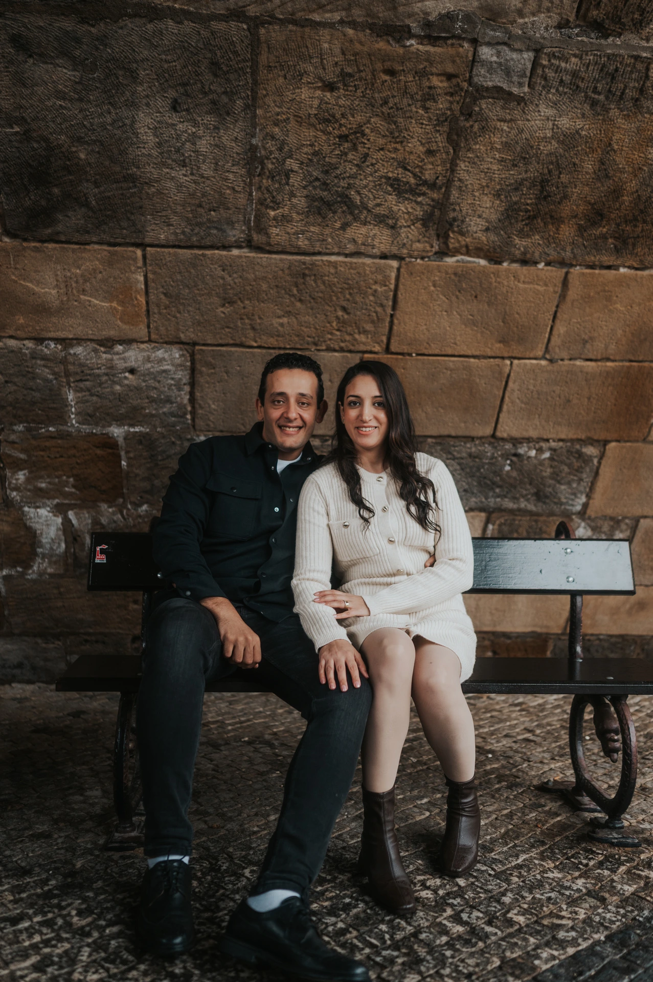 Parents sitting together and smiling towards the camera from the bench beneath Charles Bridge during their Prague family photoshoot.