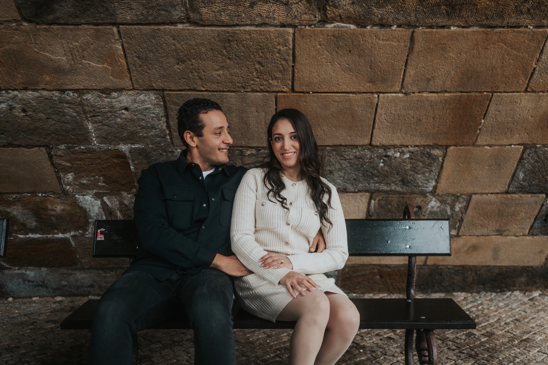 Wide portrait of the parents looking at each other while seated below Charles Bridge during their Prague family photoshoot.