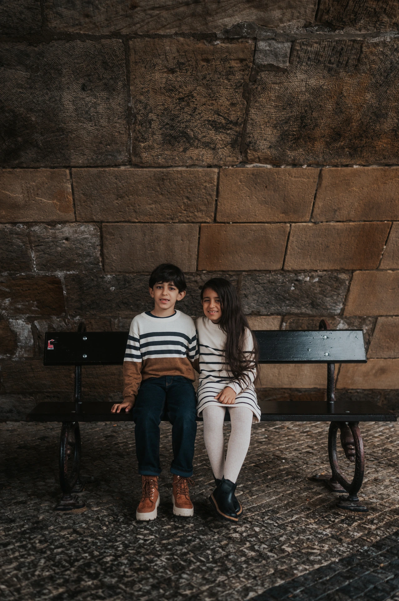 Brother and sister smiling towards the camera from a bench beneath Charles Bridge during their Prague family photoshoot.