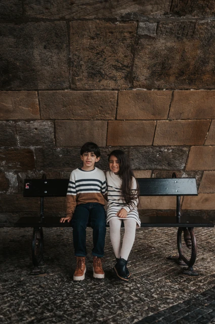 Prague family photoshoot: two smiles on the bench below Charles Bridge Brother and sister smiling towards the camera from a bench beneath Charles Bridge during their Prague family photoshoot.