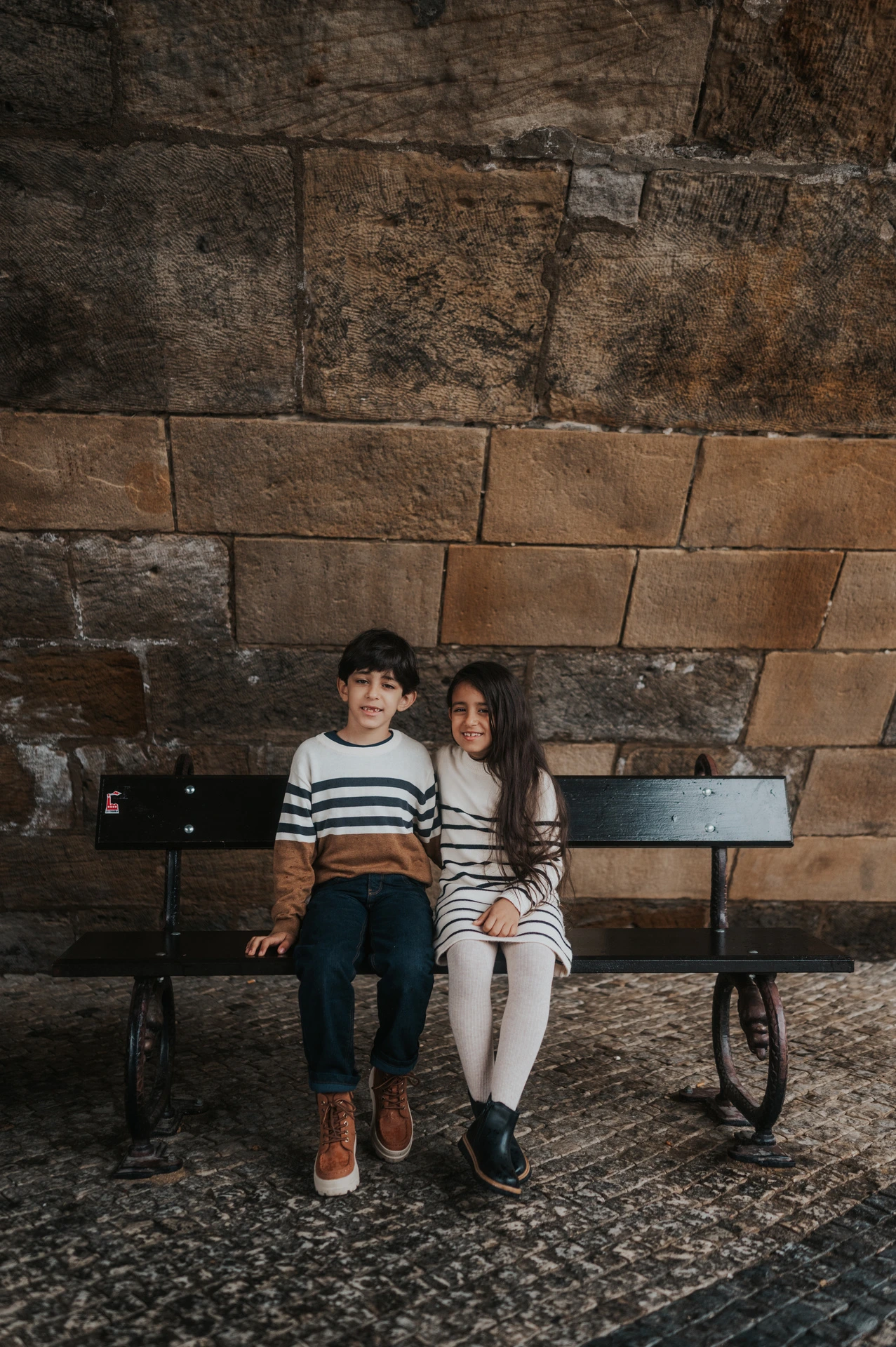 The twins sitting close together on a bench beneath Charles Bridge during their Prague family photoshoot.