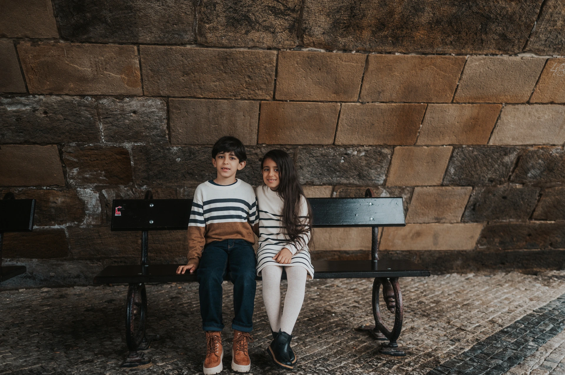 Wide photograph of the twins sitting side by side on a bench against the bridge's stone wall during their Prague family photoshoot.