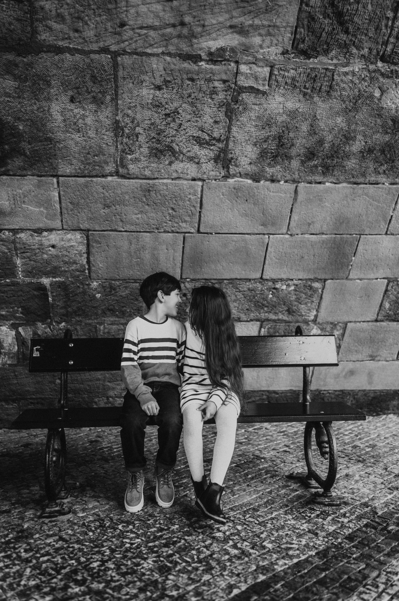 Black-and-white portrait of the twins turning towards each other on a sheltered bench during their Prague family photoshoot.
