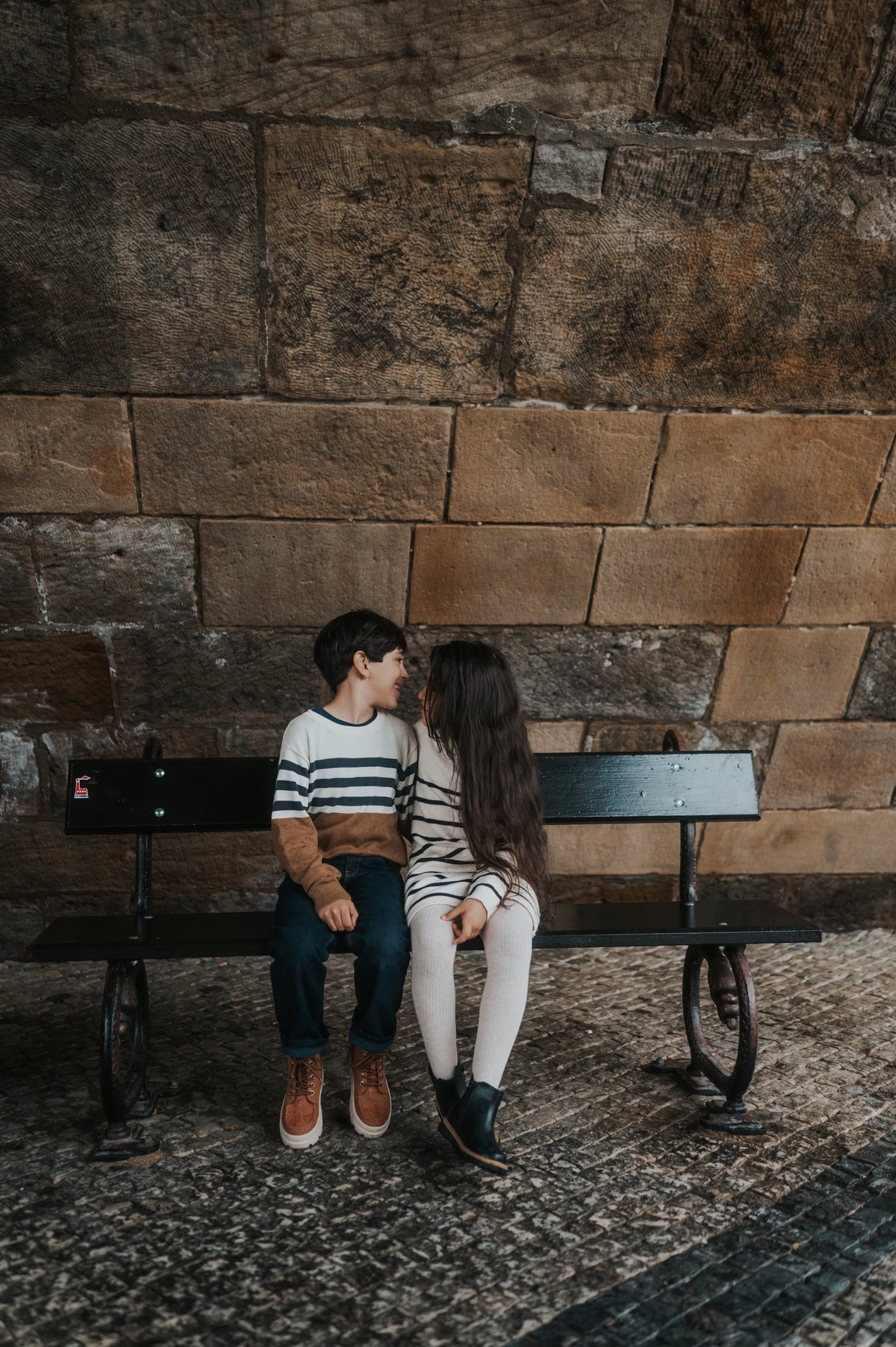The twins turning towards each other while sitting on a bench beneath Charles Bridge during their Prague family photoshoot.