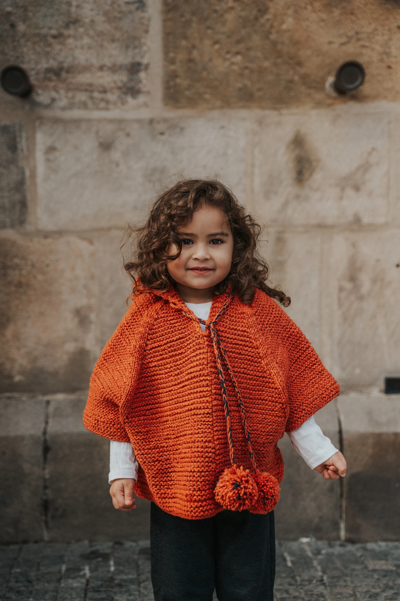 Little girl smiling in an orange knitted cape beside the stone wall during a mother and daughter family photoshoot in Prague.