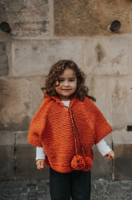 Prague family photoshoot: little girl smiling in an orange knitted cape beside the stone wall. Little girl smiling in an orange knitted cape beside the stone wall during a mother and daughter family photoshoot in Prague.