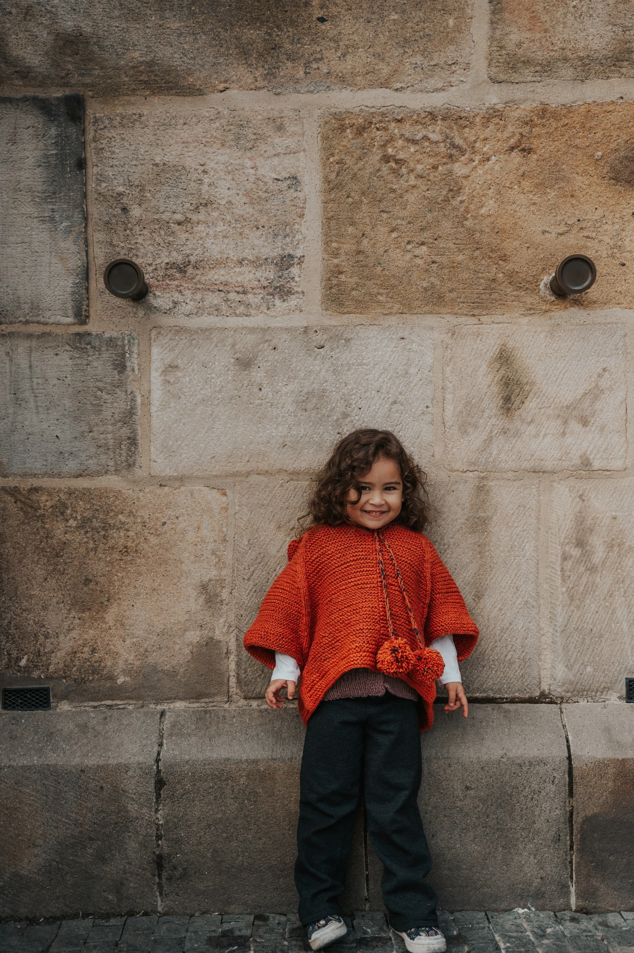 Little girl standing against a weathered stone wall during a mother and daughter family photoshoot in Prague.