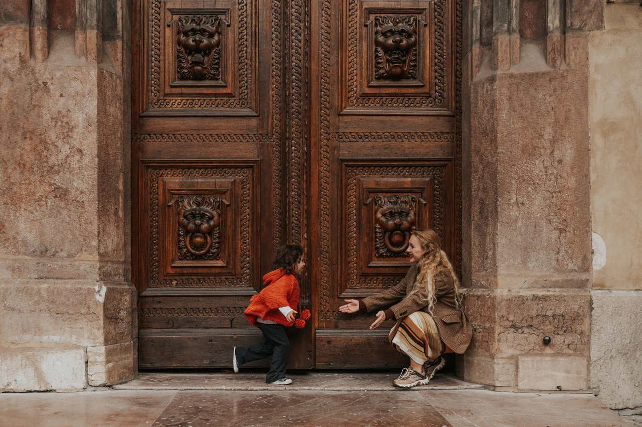 Prague family photoshoot: mother crouching beside her daughter in front of the carved doors. Mother crouching beside her daughter in front of the carved doors during a mother and daughter family photoshoot in Prague.
