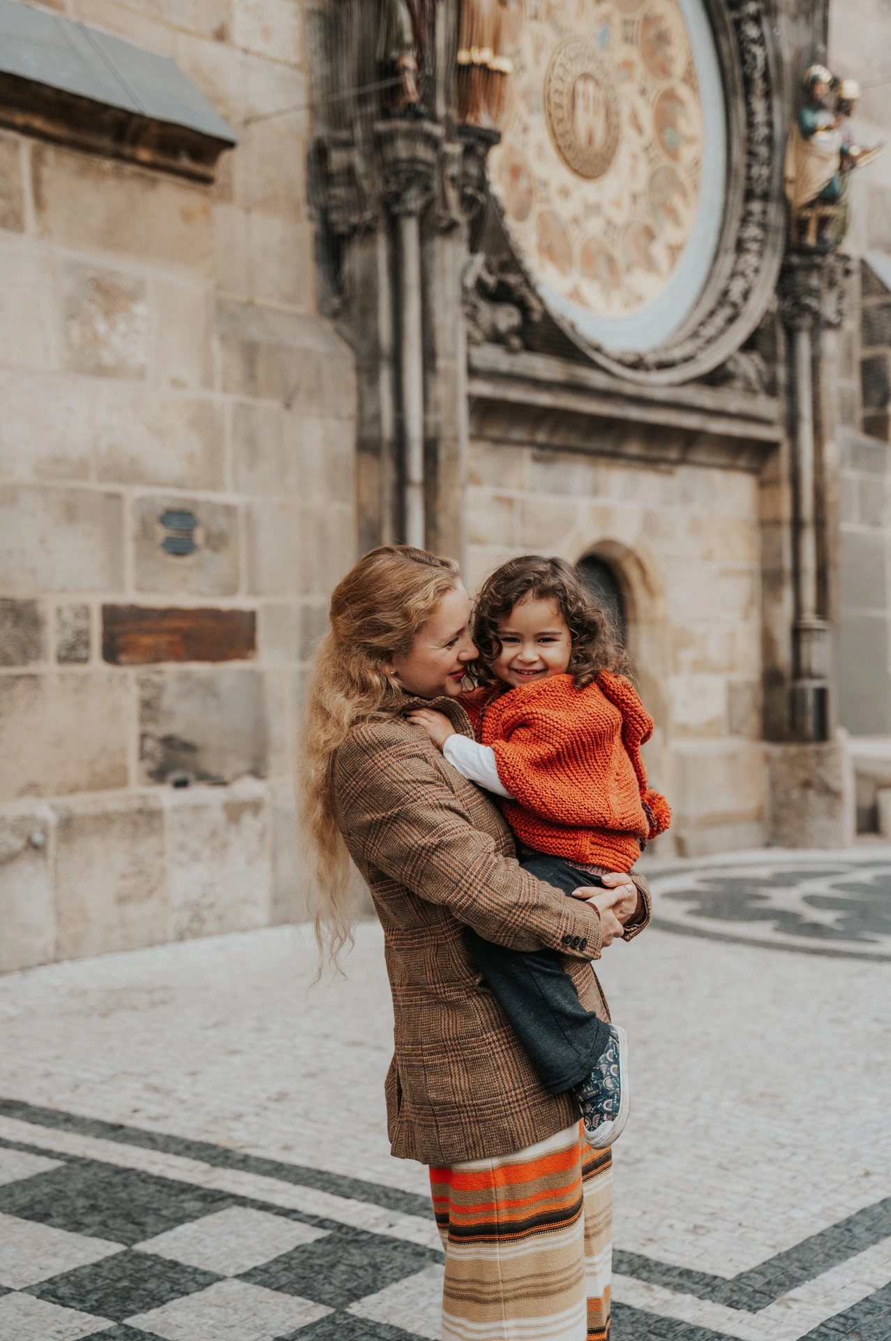 Mother smiling while holding her daughter near the clock during a mother and daughter family photoshoot in Prague.