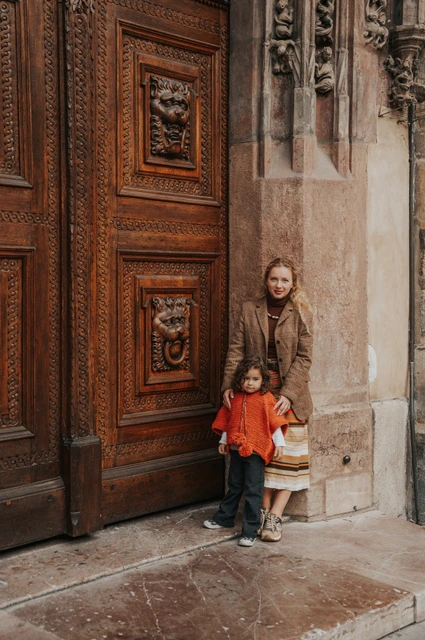 Prague family photoshoot: mother standing behind her daughter at the large wooden entrance. Mother standing behind her daughter at the large wooden entrance during a mother and daughter family photoshoot in Prague.