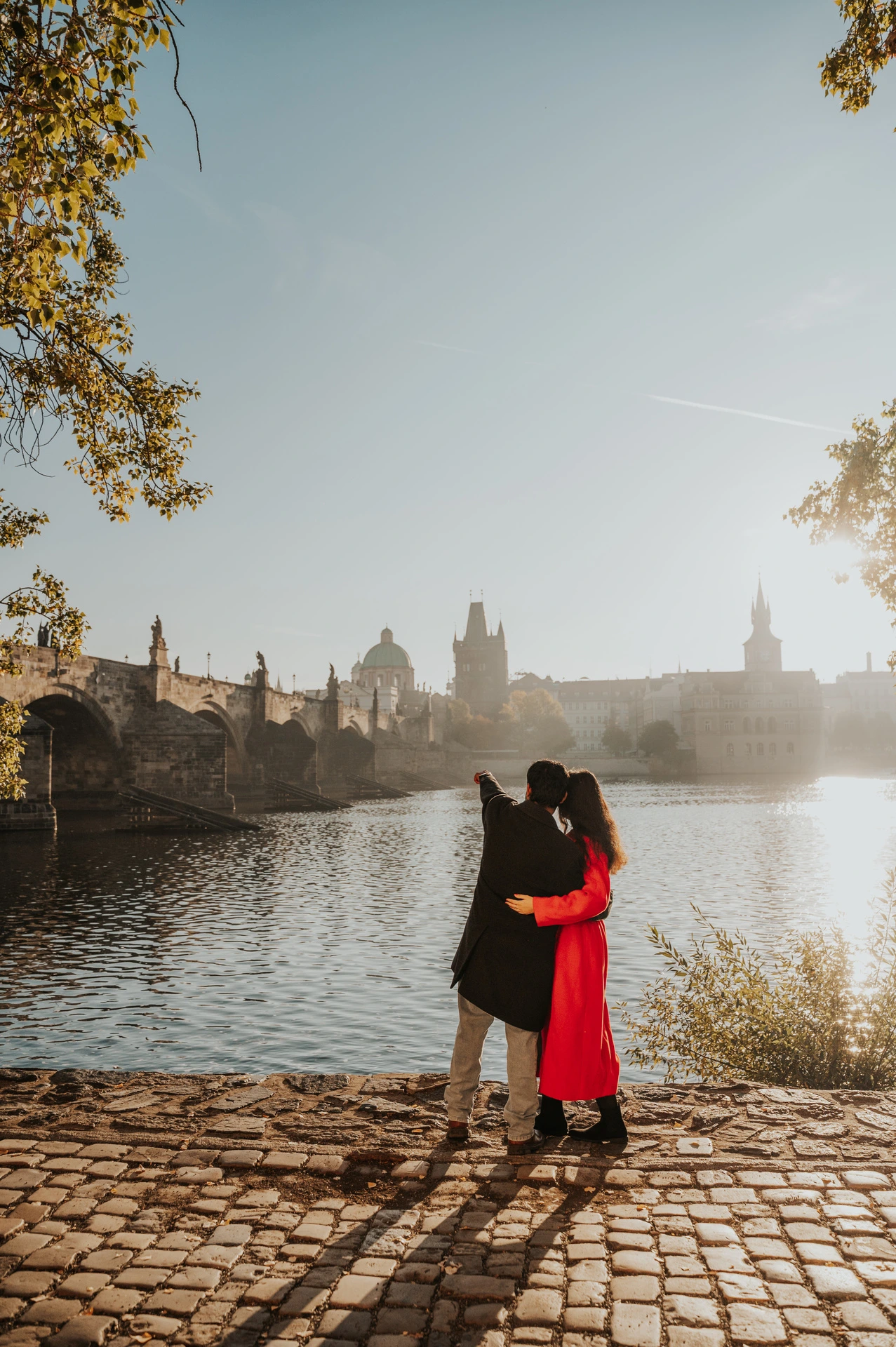Couple embracing beneath autumn leaves on the riverbank during a couple photoshoot in Prague.