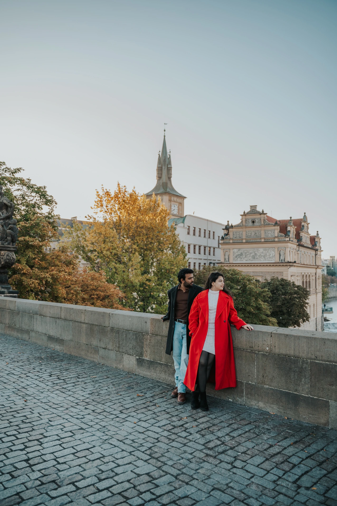 Couple leaning together beside autumn trees on Charles Bridge during a couple photoshoot in Prague.