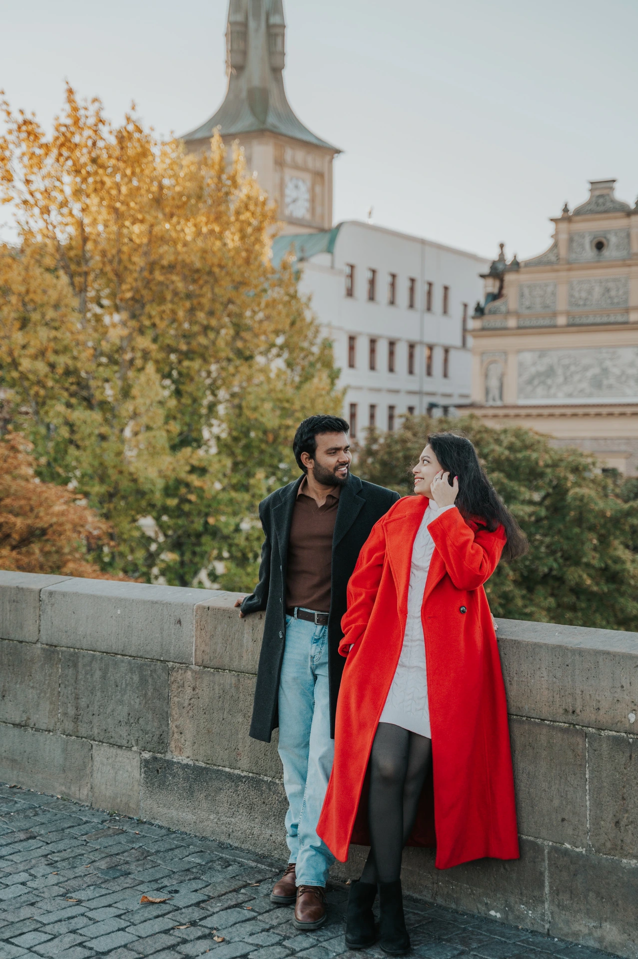 Couple smiling beside the bridge parapet in their winter coats during a couple photoshoot in Prague.