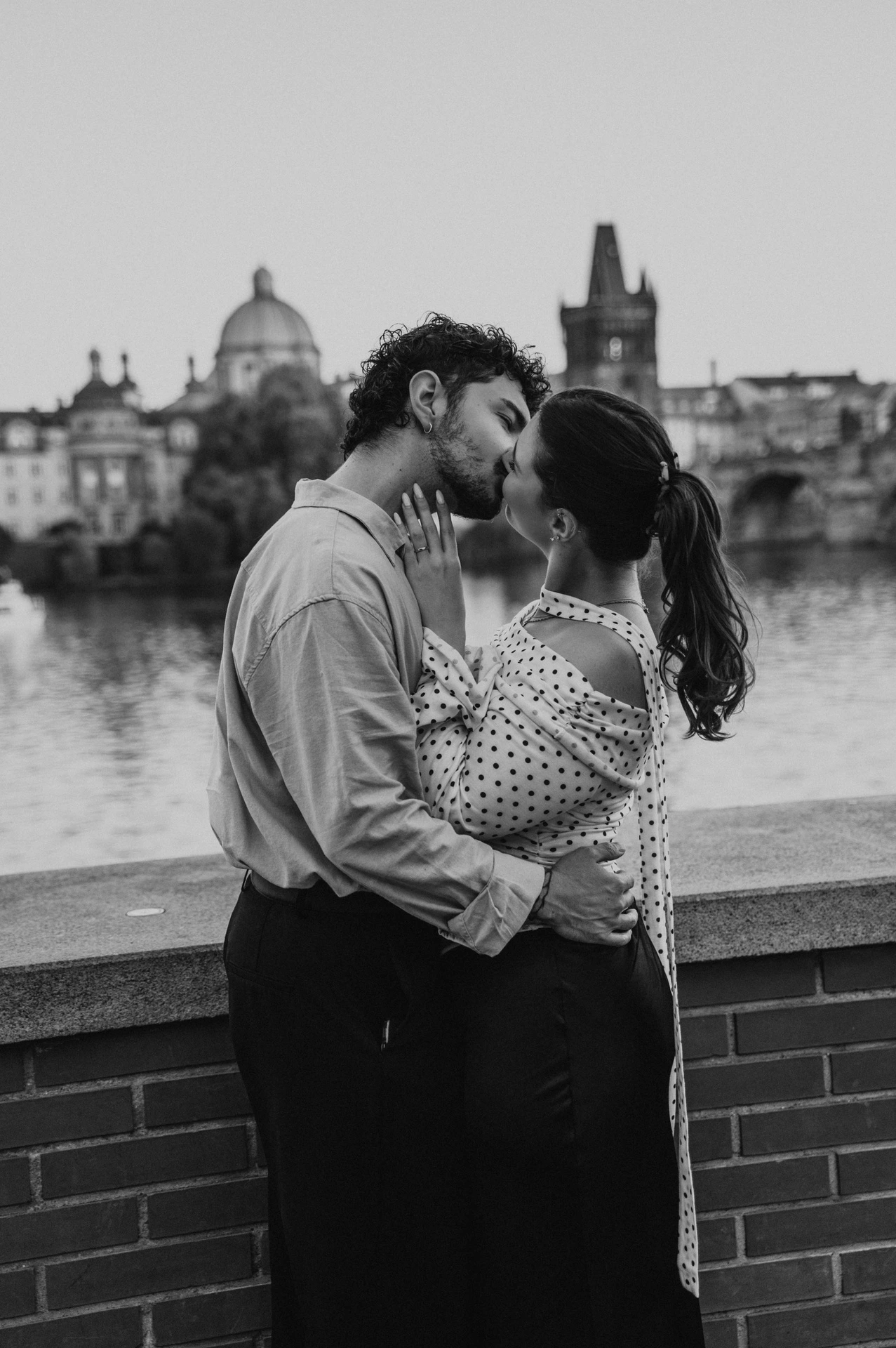Black and white kiss beside the low brick river wall during a couple photoshoot in Prague.