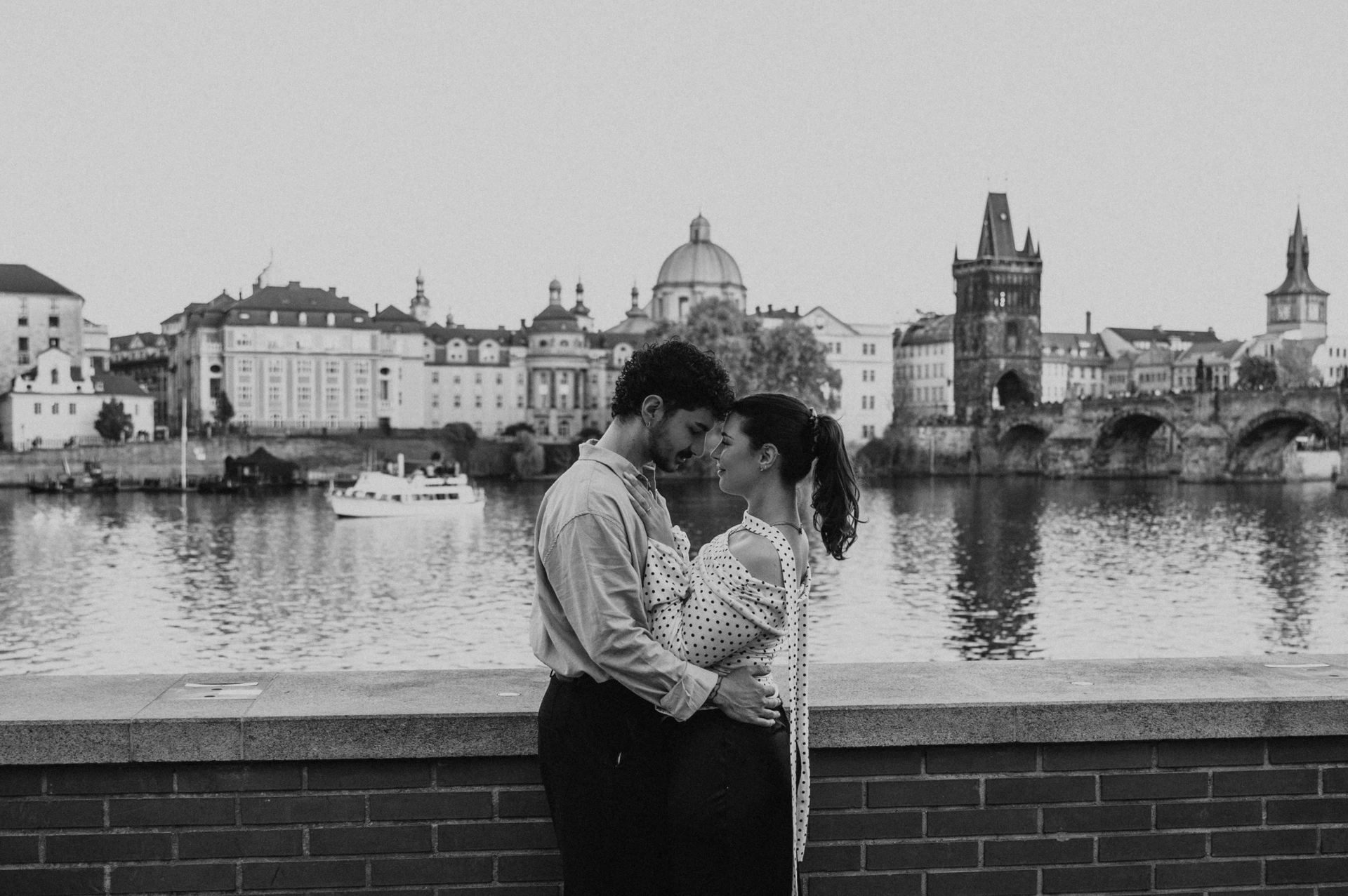 Black and white landscape portrait of a riverside kiss during a couple photoshoot in Prague.