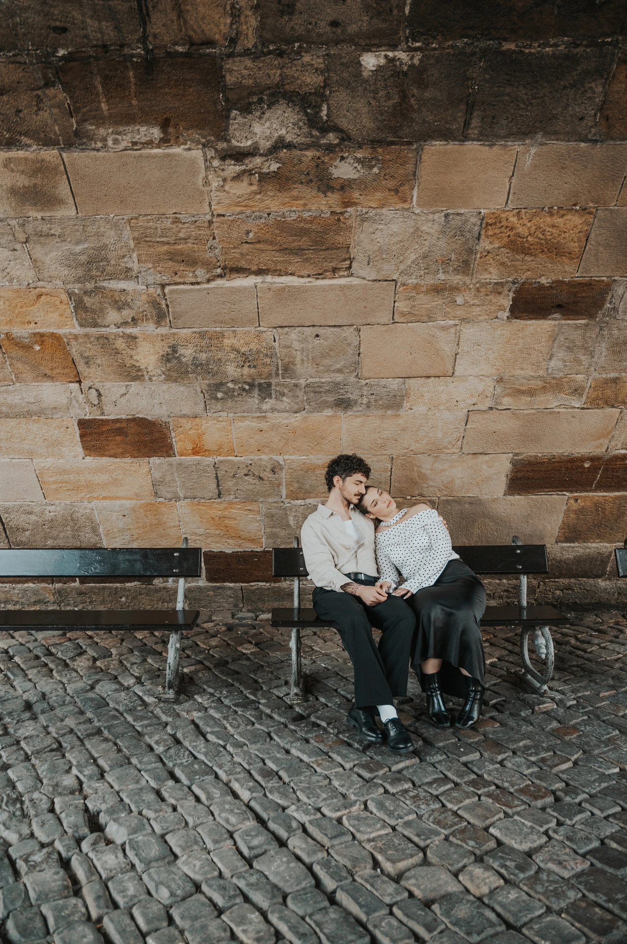 Couple cuddling on a bench against the stone bridge wall during a couple photoshoot in Prague.