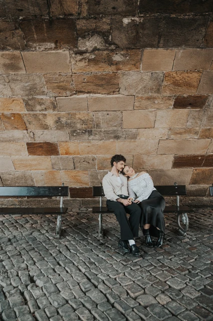 Prague couple photoshoot: couple cuddling on a bench against the stone bridge wall. Couple cuddling on a bench against the stone bridge wall during a couple photoshoot in Prague.