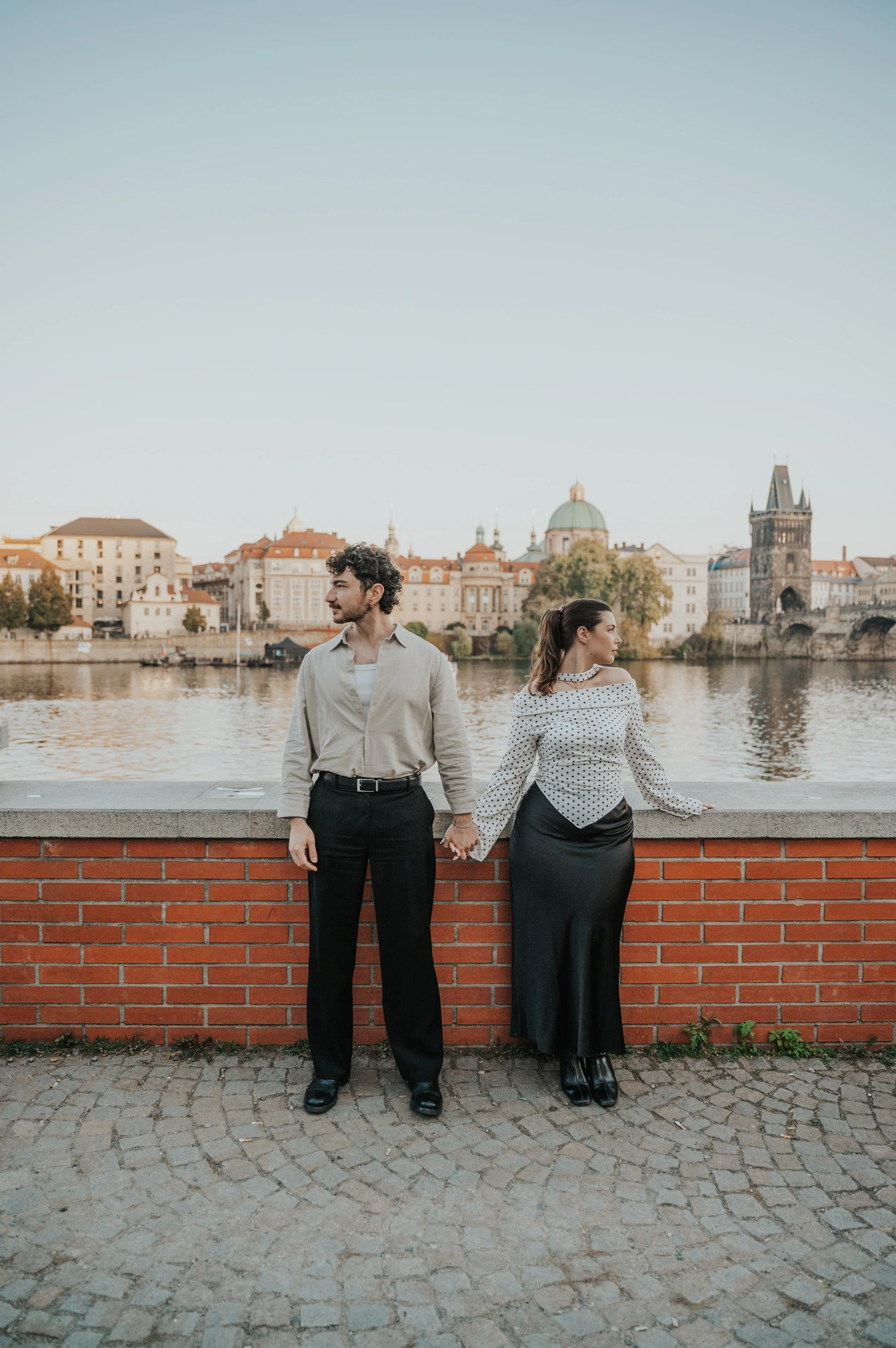 Couple holding hands against the low brick river wall during a couple photoshoot in Prague.