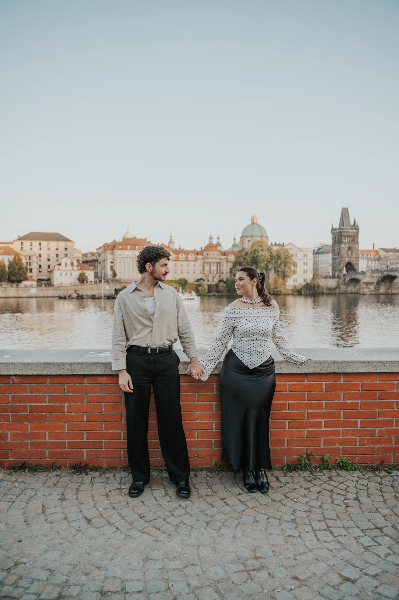 Couple looking at each other in a wide riverside portrait during a couple photoshoot in Prague.