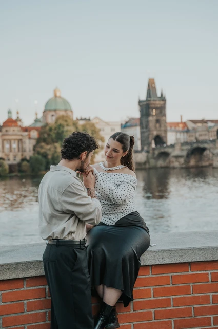 Prague couple photoshoot: couple sitting on a red riverside wall with Charles Bridge behind them. Couple sitting on a red riverside wall with Charles Bridge behind them during a couple photoshoot in Prague.