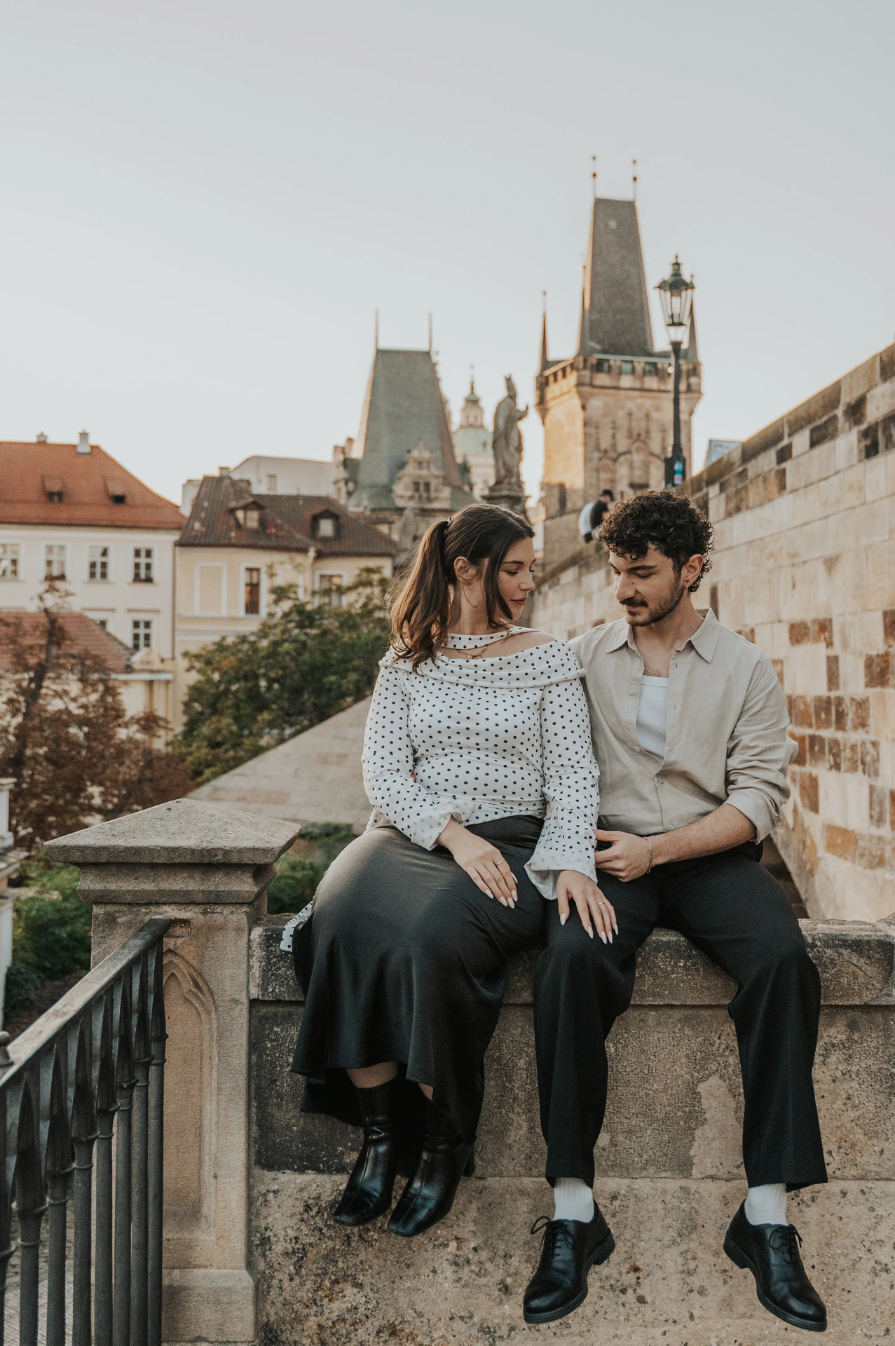 Couple sitting together on the stone railing by Charles Bridge during a couple photoshoot in Prague.
