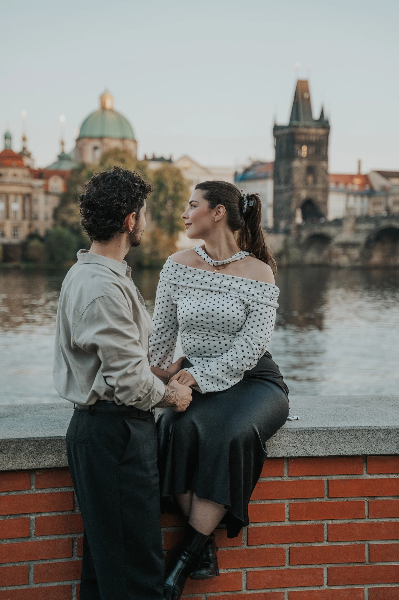 Couple smiling at one another while sitting on the riverside wall during a couple photoshoot in Prague.