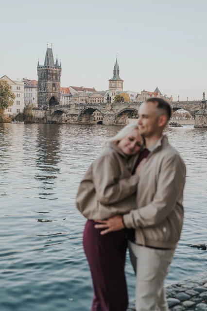 Prague couple photoshoot: couple embracing beside the Vltava with bridge towers behind them. Couple embracing beside the Vltava with bridge towers behind them during a couple photoshoot in Prague.